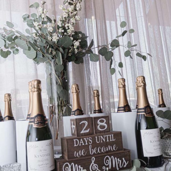 Champagne bottles surround a floral arrangement and wooden wedding countdown sign reading “428 days until we become Mr. & Mrs.”