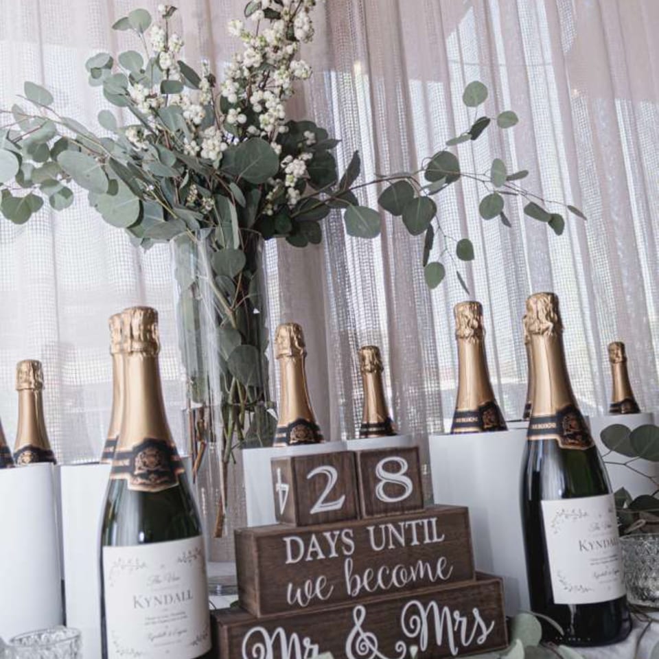 Champagne bottles surround a floral arrangement and wooden wedding countdown sign reading “428 days until we become Mr. & Mrs.”