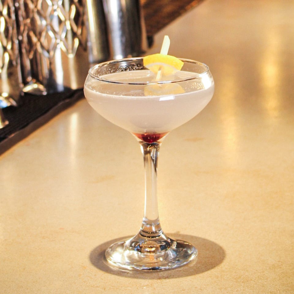Pale cocktail in a coupe glass garnished with a lemon wedge, displayed on a warmly lit restaurant bar.