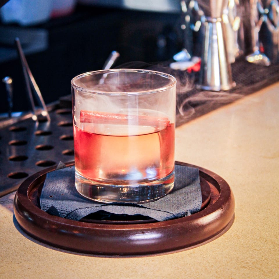 Clear tumbler of pink cocktail on a slate coaster and wooden tray at a warmly lit bar.