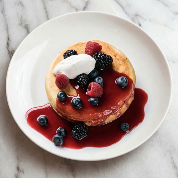 Fluffy pancake topped with whipped cream, raspberries, blueberries, and blackberries, drizzled with berry sauce.