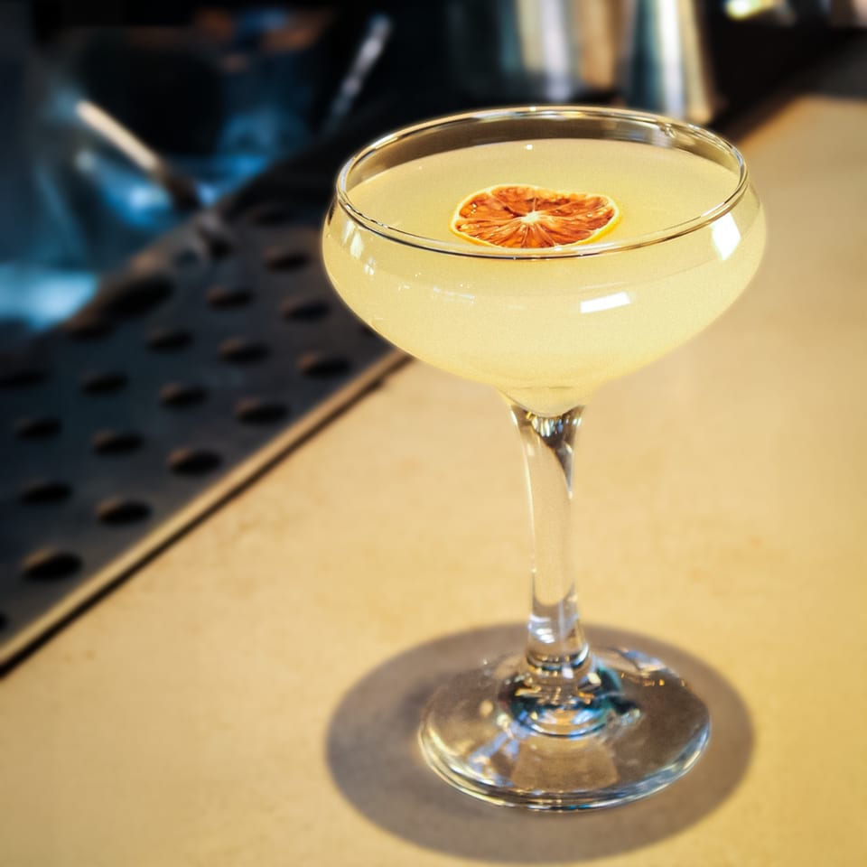 Pale yellow cocktail in a stemmed glass garnished with a dried citrus slice on a restaurant bar.