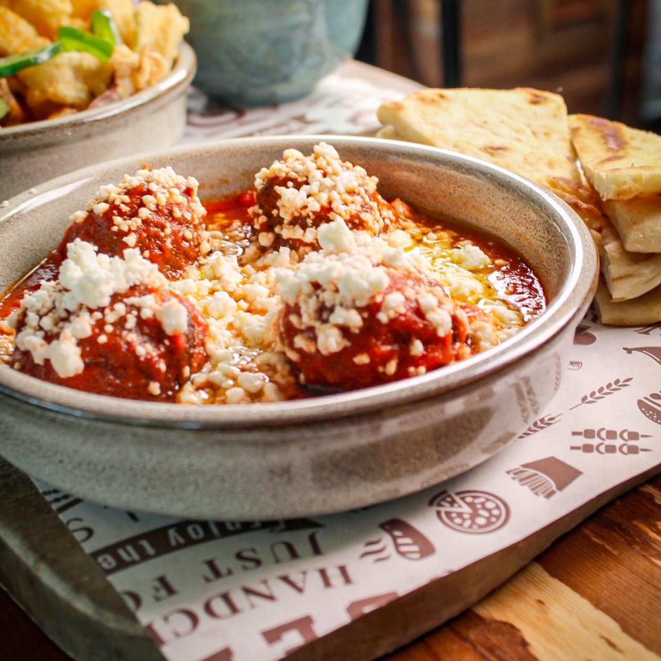 Meatballs smothered in tomato sauce and crumbled cheese, served in a metal dish with flatbread and side salad.