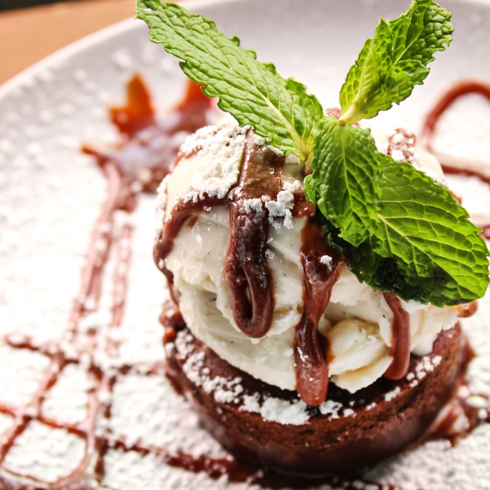 Chocolate dessert topped with vanilla ice cream, chocolate sauce, powdered sugar, and fresh mint leaves on a white plate.