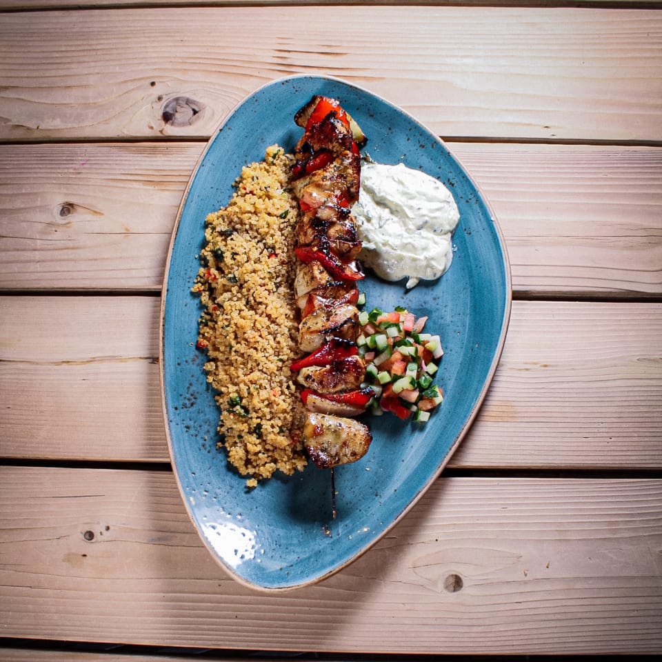 Blue plate holding grilled chicken and vegetables, quinoa, creamy tzatziki, and chopped salad on a wooden table.