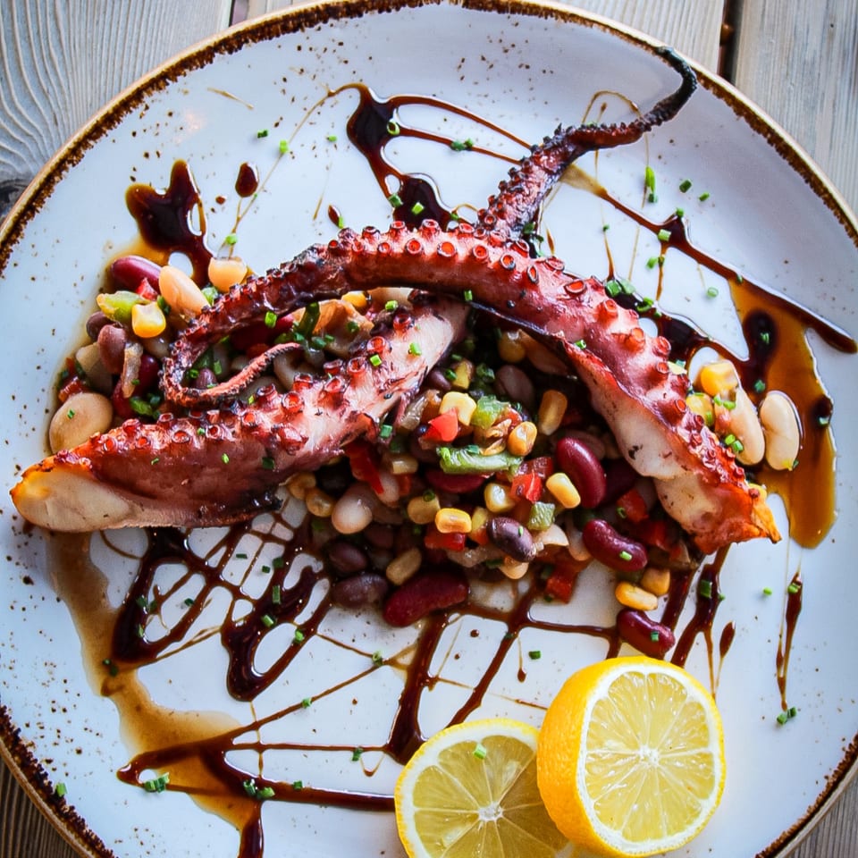 Grilled octopus tentacles served over mixed beans and vegetables, garnished with herbs, citrus slices, and drizzled sauce.