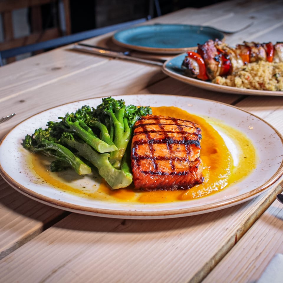 Grilled salmon fillet with char marks, broccolini, and orange sauce on a ceramic plate at a wooden restaurant table.