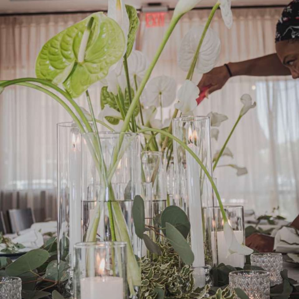 Lush white and green floral centerpiece with lit candles decorates a restaurant dining table as a person arranges flowers.
