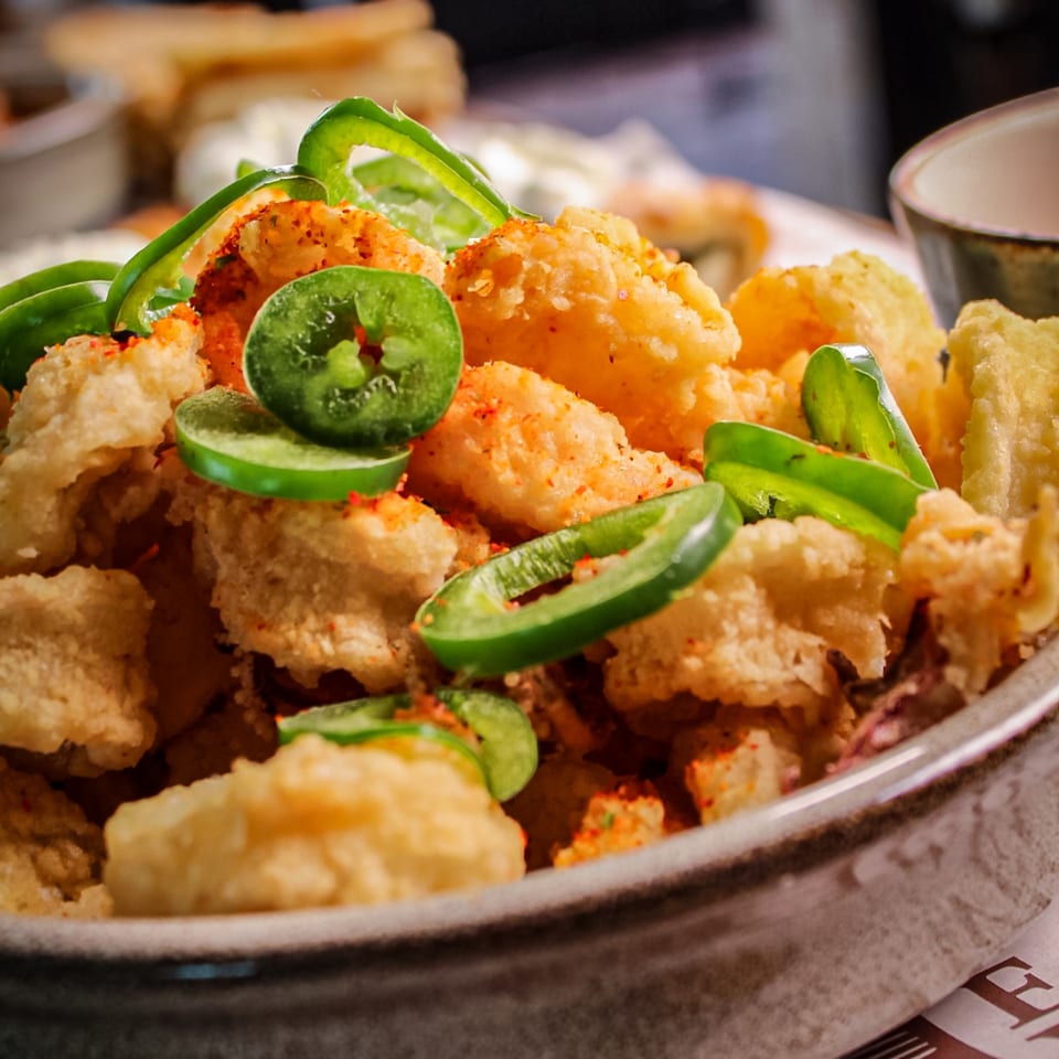 Crispy golden fried bites topped with sliced green jalapeños and red seasoning on a restaurant plate.