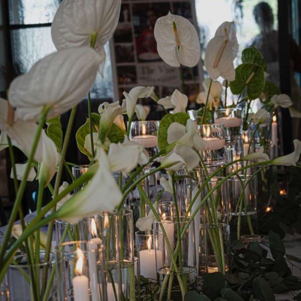 White anthurium flowers and green leaves surround glowing candles in glass holders, creating an elegant restaurant centerpiece.