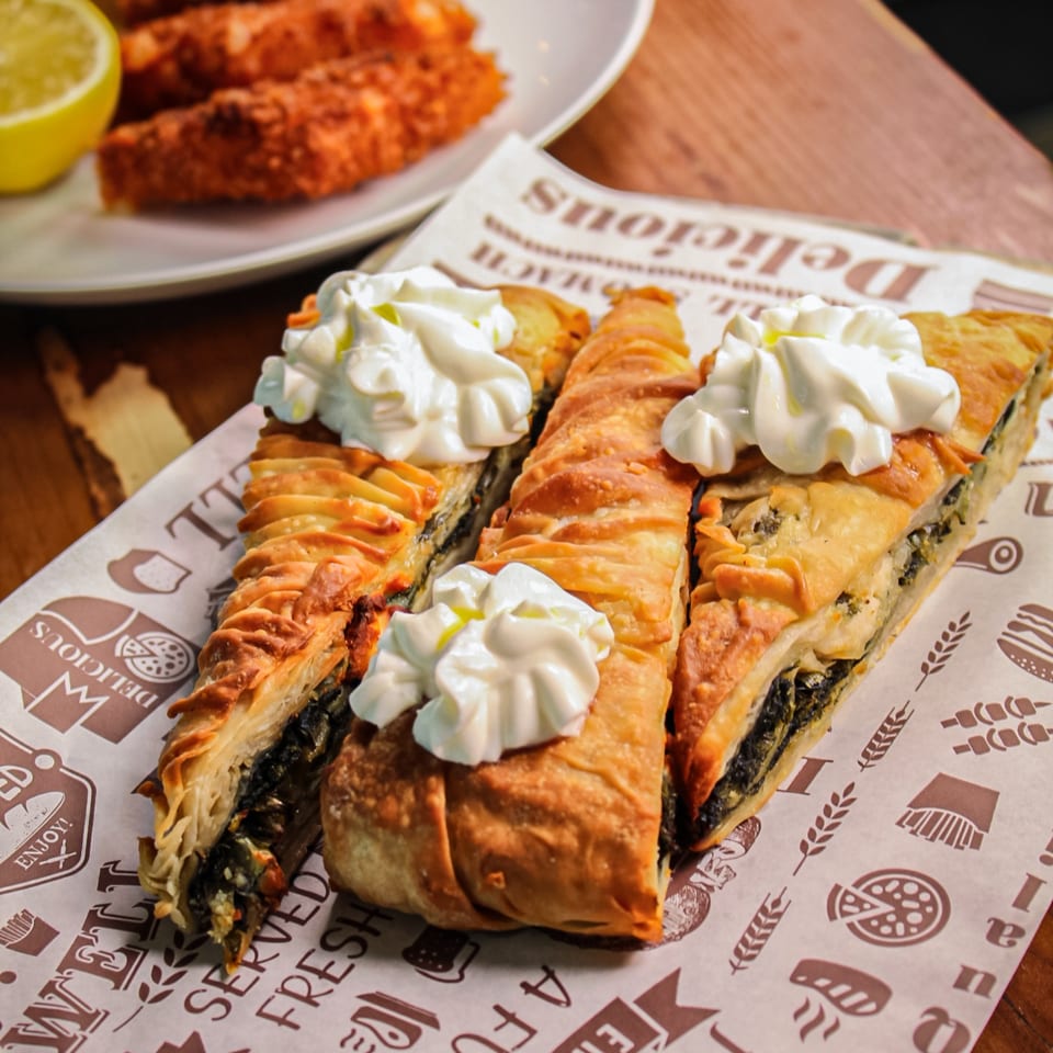 Three golden-brown filled pastries topped with whipped cream sit on branded paper beside a plate of fried food and dipping sauce.