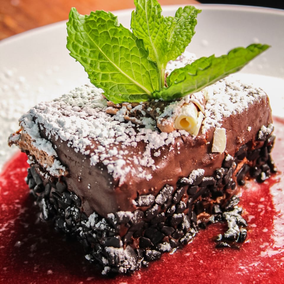 Chocolate cake coated with chocolate chips and powdered sugar, topped with mint leaves and a rolled wafer on a red plate.