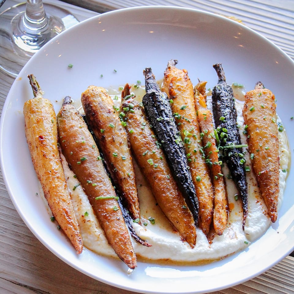 Roasted carrots in assorted colors arranged over creamy sauce and garnished with chopped chives on a white plate.