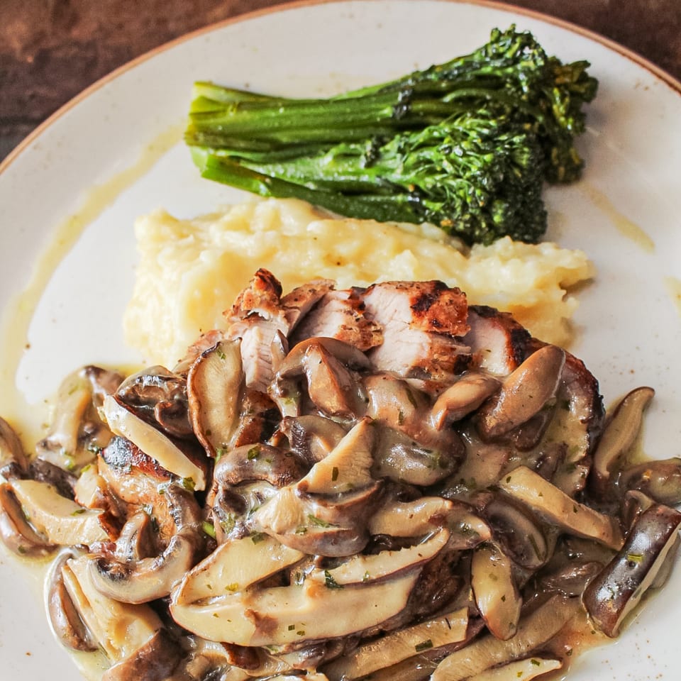 Grilled chicken topped with sautéed mushrooms, served with mashed potatoes and green vegetables on a white plate.